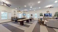 This single-floor community clubhouse is thoughtfully designed to accommodate a variety of social and recreational activities. The open-plan layout is divided into distinct functional zones, creating a versatile and welcoming environment throughout.

The game room is a central highlight, featuring a full-size pool table and a shuffleboard table positioned on a geometric patterned carpet. Multiple wall-mounted flat-screen televisions are strategically placed throughout, while pendant lighting above the game tables and ceiling fans provide both practical illumination and comfort. Bar-height tables with stools offer casual seating alongside the games area, and a dedicated cue rack keeps equipment organized.

Adjacent to the game room, the lounge area offers comfortable sectional sofas and accent chairs arranged around a large television and coffee tables. Coastal-inspired décor, including nature-themed artwork and decorative plants, contributes to a relaxed ambiance. Natural light flows generously through numerous white-shuttered windows.

The kitchen area is well-appointed with dark espresso cabinetry, granite-style countertops, wood-look vinyl flooring, and stainless steel appliances including a refrigerator and microwave. A large central dining table with seating provides an ideal space for group gatherings. Glass-panel entry doors connect this area seamlessly to the exterior.