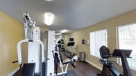 This single-level amenity space offers a comprehensive set of community features designed to support both fitness and leisure. The fitness center is well-appointed with a multi-station cable machine by Hoist, treadmills, a rowing machine, and a stationary bike, all set on wood-look flooring beneath a textured ceiling with bright overhead lighting. A window with vertical blinds allows natural light to filter into the room, creating a comfortable workout environment.

The outdoor areas are a highlight of this space. A generously sized swimming pool with gleaming turquoise water is surrounded by a clean brick-trimmed deck, lounge chairs, and mature trees that provide natural shade. Poolside seating includes round tables with red market umbrellas and rattan chairs, offering a relaxed atmosphere for outdoor gatherings under open blue skies.

Adjacent to the pool, a shaded pergola area provides a covered retreat with lounge seating and potted greenery, framed by a neatly trimmed hedge for privacy. A dedicated BBQ patio features multiple freestanding gas grills beneath a covered overhang, with café-style outdoor furniture set on a paved surface bordered by a manicured lawn. Together, these amenities create a vibrant, resort-like outdoor living experience.