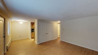 This single-floor space is designed with an open and functional layout that maximizes usability. The main living area features smooth hardwood-style flooring in a warm honey tone, providing a clean and cohesive look throughout. Neutral white walls and a textured ceiling contribute to a bright, airy atmosphere, while flush-mounted ceiling lights ensure consistent illumination across the space.

A set of classic double doors with arched paneling is centrally positioned along one wall, offering generous built-in closet storage that is both practical and visually integrated into the room. Adjacent to the main living area, a partially visible kitchen features light wood cabinetry, dark countertops, and track lighting overhead, offering a compact yet well-appointed cooking space.

A hallway visible on the right side of the space leads further into the unit, suggesting additional rooms or utility areas beyond. The overall design is clean and understated, with white baseboards framing the flooring and adding a polished finishing touch. The space presents a straightforward, move-in-ready environment with a neutral palette suited to a variety of furnishing styles.