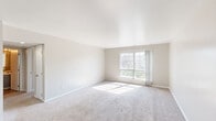 This single-floor space offers a clean, neutral interior designed for comfort and practicality. The living area is generously proportioned, featuring soft wall-to-wall carpeting, light beige walls, and a large multi-pane window that floods the room with natural light and offers views of surrounding greenery. The open, unobstructed layout provides flexible arrangement possibilities.

The bedroom is well-sized with matching carpet flooring and its own window fitted with blinds for adjustable light control. It includes a built-in closet with upper shelving and a hanging rod, offering solid storage capacity. An additional carpeted room provides versatile space suitable as a second bedroom, office, or flex room, also benefiting from natural light.

The kitchen is compact and efficiently laid out, featuring gray flat-front cabinetry on both upper and lower sections, light countertops, a white tile backsplash, and wood-look vinyl plank flooring. A full-size refrigerator is included. The bathroom features classic floor-to-ceiling square tile in a warm taupe tone surrounding a soaking tub with overhead shower, complemented by a large wall mirror and updated fixtures. Throughout the space, the consistent neutral palette and abundant natural light create a bright, cohesive atmosphere.