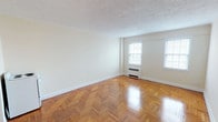 This single-floor unit showcases a consistent design aesthetic throughout, anchored by gleaming herringbone-pattern parquet wood flooring that extends across the main living areas. The living room is generously proportioned, featuring cream-toned walls, a textured ceiling, and multiple windows that admit a steady supply of natural light. A wall-mounted radiator unit provides heating, and clean white baseboards frame the perimeter of each room.

A second room, likely used as a bedroom, mirrors the same herringbone flooring and neutral palette, with two windows fitted with blinds positioned side by side, allowing for ample daylight. A radiator sits beneath the windows, consistent with the building's heating system.

The kitchen is compact and efficiently laid out, featuring white cabinetry, a white range, and a sink with straightforward counter space. It opens directly to the main living area through an arched passageway, creating a connected feel between the spaces.

The bathroom includes a full-size soaking tub with an overhead shower and chrome fixtures. The walls are clad in cream-colored tiles accented by a contrasting black tile border, lending a classic, retro character to the space. Overall, the unit presents a clean, functional layout with timeless design details.