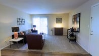 This single-floor residence features a well-proportioned living room that serves as the centerpiece of the space. The room is finished with a combination of hardwood flooring near the entryway and wall-to-wall carpeting throughout the main seating area, creating a comfortable and functional layout. A large sliding glass door dominates one wall, fitted with vertical blinds that allow for adjustable natural light and provide access to an outdoor balcony. The generous amount of natural light filtering through the door gives the room an open, airy feel. The neutral, light-toned walls provide a versatile backdrop that complements a variety of furnishing styles. The living area comfortably accommodates a sectional sofa, an armchair, and a coffee table, demonstrating its practical use of space. Additional storage and display options are provided by a dark wood credenza and an open media console positioned along the adjacent wall. Warm accent lighting enhances the room's welcoming atmosphere, making it a functional and inviting area for everyday living.
