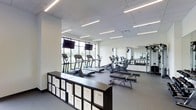 This contemporary fitness center is thoughtfully designed to support a full range of workout activities within a clean, open layout. The space features a sleek, neutral color palette of whites and grays, complemented by a drop-tile ceiling with rows of linear LED lighting that ensures bright, even illumination throughout.

Along the perimeter, large floor-to-ceiling windows invite abundant natural light and offer refreshing views of the surrounding greenery, creating an energizing atmosphere for cardio workouts. A lineup of modern cardio machines — including treadmills, ellipticals, and a stationary bike — is positioned to take full advantage of these views, and dual flat-screen monitors mounted above the windows provide entertainment options during exercise.

Full-length mirrors line one wall, visually expanding the space while allowing users to monitor their form during strength training. A dedicated weight and resistance training area is visible in the mirror's reflection, suggesting a comprehensive equipment offering. The flooring is a durable gray surface suited for high-traffic athletic use.

A practical storage unit with labeled white bins runs along one side of the room, offering organized storage for accessories and personal items. The overall design strikes a balance between functionality and modern aesthetics, making this fitness center a well-appointed and inviting space for daily exercise.