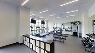 This contemporary fitness center is thoughtfully designed to support a full range of workout activities within a clean, open layout. The space features a sleek, neutral color palette of whites and grays, complemented by a drop-tile ceiling with rows of linear LED lighting that ensures bright, even illumination throughout.

Along the perimeter, large floor-to-ceiling windows invite abundant natural light and offer refreshing views of the surrounding greenery, creating an energizing atmosphere for cardio workouts. A lineup of modern cardio machines — including treadmills, ellipticals, and a stationary bike — is positioned to take full advantage of these views, and dual flat-screen monitors mounted above the windows provide entertainment options during exercise.

Full-length mirrors line one wall, visually expanding the space while allowing users to monitor their form during strength training. A dedicated weight and resistance training area is visible in the mirror's reflection, suggesting a comprehensive equipment offering. The flooring is a durable gray surface suited for high-traffic athletic use.

A practical storage unit with labeled white bins runs along one side of the room, offering organized storage for accessories and personal items. The overall design strikes a balance between functionality and modern aesthetics, making this fitness center a well-appointed and inviting space for daily exercise.