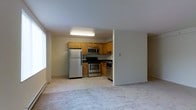 This single-floor space features an open-concept design that connects the kitchen and living area seamlessly. The kitchen is equipped with stainless steel appliances, including a refrigerator, gas range, and over-the-range microwave. Warm-toned wooden cabinetry lines the walls, providing ample storage, while a sink with an overhead faucet is positioned along the far counter. The kitchen flooring is light-toned tile, which transitions into plush carpeting in the adjacent living area. Neutral beige walls and a white ceiling keep the space feeling bright and airy. Fluorescent overhead lighting illuminates the kitchen, while natural light filters in through a window visible on the left side of the living area. A baseboard heating unit runs along the perimeter, offering an efficient heating solution. The open layout between the kitchen and living space creates a functional and cohesive environment suitable for everyday living.