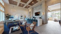 This single-story amenity space is designed in a coastal contemporary style, blending natural materials with refined finishes throughout its generously proportioned interior. The main lounge area commands attention with soaring ceilings, floor-to-ceiling windows, and patterned drapery that frames sweeping views of palm-lined outdoor surroundings, flooding the space with abundant natural light. Neutral upholstered seating, navy blue area rugs, and curated coastal artwork create a welcoming, resort-like atmosphere.

Adjacent to the lounge, a bar area features a stacked stone accent wall, a wall-mounted flat-screen television, and counter-height seating along a sleek white bar top — ideal for social gatherings. A coffered ceiling with warm wood inlays and recessed lighting adds architectural distinction to this zone.

A game area includes a billiard table set on light hardwood flooring, complemented by a built-in banquette with reclaimed wood backdrop and pendant lighting, offering an inviting nook for casual socializing.

Two dedicated office or meeting rooms provide functional workspace, each featuring clerestory or tall windows, woven pendant light fixtures, and access to natural light. One room incorporates a textured accent wall and an L-shaped desk arrangement.

Throughout, large-format tile flooring, stone-clad columns, and thoughtfully layered lighting unify the space with a polished, coastal aesthetic.