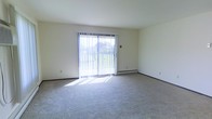 This single-floor residential space offers a clean, functional layout designed for comfortable everyday living. The living area is a standout feature, finished with wall-to-wall carpeting that adds warmth and softness underfoot. A large sliding glass door serves as the room's focal point, fitted with vertical blinds and opening to an outdoor balcony, allowing generous amounts of natural light to fill the interior. The baseboard heating system running along the exterior wall ensures year-round comfort. The walls are finished in a neutral, light tone that enhances the bright and airy atmosphere of the space. Dark baseboard trim provides a subtle contrast to the light flooring and walls, adding a touch of definition to the room's clean lines. The overall ambiance is calm and understated, offering a versatile blank canvas well-suited for a variety of interior arrangements and décor styles. The single-floor layout provides convenient, step-free living throughout the entire space.