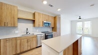 This well-designed single-story unit offers a thoughtfully organized layout across a comfortable footprint. The open-concept kitchen and living area serves as the heart of the space, featuring light wood-grain cabinetry extending from floor to ceiling, a full suite of stainless steel appliances including a refrigerator, range, dishwasher, and built-in microwave. A tiled backsplash adds texture, while a central island with a crisp white countertop provides additional workspace and casual seating. Light wood-toned flooring runs throughout the main living areas, complementing the bright, neutral color palette. French doors in the living area introduce abundant natural light and open to an outdoor balcony with green views. A wide hallway connects the main living space to the private bedroom, which is generously sized, carpeted for comfort, and includes a ceiling fan and two windows with outdoor views. Two closets provide ample storage. The bathroom features a double-sink vanity with warm wood cabinetry, white quartz countertops, matte black fixtures, and a walk-in shower with wood-look tile. Clean lines and modern finishes define the overall aesthetic throughout.