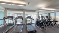 The Upstairs Fitness Center is a well-appointed exercise facility designed to support an active lifestyle. The workout area features a clean, modern aesthetic with a crisp white ceiling accented by sleek linear lighting fixtures that provide bright, even illumination throughout the space. Durable rubber flooring lines the exercise floor, providing a safe and functional surface for workouts. The cardio section is equipped with multiple Cybex treadmills and elliptical machines, arranged to face a bank of large windows that flood the room with natural light. Full-length mirrors along the walls enhance the sense of openness and allow exercisers to monitor their form. The windows offer elevated views of the outdoor pool area below, where a generously sized swimming pool is surrounded by a spacious sundeck lined with lounge chairs and patio umbrellas. The pool area is framed by mature trees and the broader residential community beyond. Access to the fitness center is provided through a covered exterior entryway featuring rustic wood-framed glass doors, decorative wall sconces, and planter accents — creating a welcoming transition between the outdoor amenity space and the gym interior. Together, these amenities form a cohesive and inviting recreational environment.