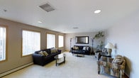 This single-floor space features a generously sized lounge area designed for comfort and functionality. The room is finished with light-colored tile flooring that extends throughout, contributing to a bright and open atmosphere. Neutral-toned walls are complemented by gold-framed windows fitted with vertical blinds, allowing for adjustable natural light intake along one side of the room.

The seating arrangement includes two dark upholstered sofas accented with patterned throw pillows, positioned around a sleek glass-topped coffee table. Additional seating is available with a patterned accent chair situated toward the far end of the room. A framed wall mirror and tall floor lamps add decorative depth to the space, while potted tropical plants introduce natural elements that soften the overall aesthetic.

The ceiling is smooth and white, featuring recessed lighting and a centrally positioned HVAC vent, ensuring even climate control throughout. Baseboard heating units line the perimeter walls, providing supplemental warmth. The layout is open and unobstructed, making the space feel expansive and easily adaptable for various uses, from casual gathering to quiet relaxation.