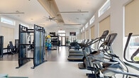 This well-appointed fitness center offers a generously sized, single-floor layout designed to accommodate a full range of workout activities. The space is defined by soaring vaulted ceilings with large ceiling fans that promote airflow throughout, while clerestory windows lining the upper walls flood the interior with natural light. Roller shades provide adjustable light control, ensuring a comfortable environment at any time of day. The durable epoxy-coated flooring runs throughout, offering both resilience and a clean, polished aesthetic.

The cardio zone features a row of treadmills, elliptical machines, stair climbers, and connected stationary bikes, including Peloton-style cycles. A dedicated strength area includes a Matrix functional trainer, a cable machine system, a squat rack with bench, and a comprehensive dumbbell rack ranging across multiple weight increments. A free-standing multi-use equipment tower with battle ropes, medicine balls, and resistance tools serves as a versatile functional training hub.

A distinctive decorative lattice partition separates a stretching and yoga alcove, furnished with a teal built-in bench and neatly organized yoga mat cubbies. Wall-mounted flat-screen TVs and full-length mirrors enhance the space. A set of black-framed glass double doors opens directly to an outdoor walkway, connecting the fitness center to the broader community.