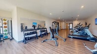 This fitness center offers a generously sized, single-floor exercise space designed to support a full range of workout routines. The room features warm-toned hardwood-style flooring throughout, recessed ceiling lighting, and crisp white walls that contribute to a clean, energizing atmosphere. Large floor-to-ceiling windows at one end of the room invite abundant natural light and provide outdoor views, enhancing the open and airy feel of the space.

The cardio section is well-stocked with multiple treadmills, elliptical trainers, stationary bikes, and rowing machines, each positioned on protective floor mats. Wall-mounted flat-screen televisions are positioned throughout the room for entertainment during workouts. The strength training area includes a multi-station cable machine, adjustable weight benches, and a full rack of dumbbells displayed on a tiered free-weight stand backed by a wall mirror. A dedicated functional training zone near the windowed wall houses medicine balls, stability balls, and resistance accessories on an organized rack.

The space is accessed through bold navy-framed glass double doors and features a water station and posted guidelines near the entrance. The layout is thoughtfully arranged to maximize usability while maintaining clear circulation paths throughout the entire fitness floor.