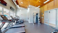 This single-floor amenity facility offers a thoughtfully designed layout that combines fitness, relaxation, and productivity across three distinct spaces connected by two hallways. The centerpiece is a generously sized exercise room featuring high, vaulted ceilings with striking warm wood paneling, exposed ductwork, and ceiling fans that lend the space an open, airy feel. The fitness area is outfitted with a comprehensive range of equipment, including treadmills, ellipticals, a cable weight machine, and free weights with a bench, all set on durable rubber flooring. Large windows along one wall flood the room with natural light and offer views of an outdoor pool area, while wall-mounted televisions provide entertainment during workouts. The living room offers a welcoming lounge atmosphere, featuring wood-look vinyl flooring, elevated ceilings with a tray detail accented by warm wood trim, and high clerestory windows that draw in natural light. Comfortable sectional seating, a large flat-screen television, and framed movie posters give the room a relaxed, social character. The office space is finished with hardwood flooring and wainscoting in a wood-tone panel design, providing a clean and professional environment with glass-door entryways and a workstation setup.