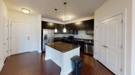 Located at 735 Ninth St in Durham, NC, this single-floor residence combines modern design with functional living spaces. The kitchen is a standout feature, showcasing rich dark cabinetry, granite countertops, a mosaic tile backsplash, and a full suite of stainless steel appliances including a French door refrigerator, electric range, microwave, and dishwasher. A central island with a granite top and pendant lighting above adds both prep space and a stylish focal point. Rich hardwood flooring flows through the kitchen and main living areas, lending warmth and continuity throughout. The living area is spacious and bright, benefiting from large windows dressed with blinds and direct access to a private balcony through glass French doors, allowing natural light to fill the room. A ceiling fan provides year-round comfort. The bedroom continues the airy feel with plush carpeting, its own ceiling fan, and additional balcony access through French doors with a transom window above. The bathroom is finished with large-format tile flooring, a dark-framed vanity with granite countertop, and a glass-enclosed shower. Clean white walls and classic panel doors tie the interior together with a polished, contemporary aesthetic throughout.