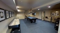 This single-floor recreation space is thoughtfully designed for entertainment and social interaction. The room is anchored by a full-size, dark-finished pool table positioned beneath a stylish pendant light fixture, creating a focal point that draws the eye immediately upon entry. A shuffleboard table lines one wall, adding a classic gaming element to the space.

The room features neutral-toned walls complemented by blue-gray carpet flooring, lending a clean, cohesive aesthetic throughout. Recessed lighting works in tandem with the overhead pendant fixture to provide balanced, ambient illumination across the space. A wall-mounted flat-screen television is positioned above a small counter or bar ledge, flanked by framed artwork, creating a media and lounge zone ideal for gatherings.

Bar-height seating is available near the media area, offering casual perching spots for spectators and guests. Multiple doors provide convenient access points throughout the space. The layout is open and well-organized, allowing comfortable movement between the various activity zones. Overall, the space presents a well-appointed recreational environment suited for hosting a variety of leisure activities.