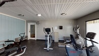 Located at 139 La Fontenay Dr in Louisville, Kentucky, this dedicated fitness facility is housed on a single floor and designed to support a comprehensive workout experience. The space features a distinctive beadboard ceiling finished in white, which lends a clean, structured aesthetic throughout the room. The walls are painted in neutral gray tones, with one accent wall finished in a soft light blue vertical paneling, adding subtle visual interest to the interior.

The facility is outfitted with a varied selection of cardio equipment, including multiple treadmills, elliptical trainers, and a stationary cycle, arranged to allow comfortable circulation between machines. A multi-station strength training unit with adjustable benches and a rowing machine occupies a dedicated area, providing options for resistance and full-body training. Rubber flooring throughout the space offers durability and safety underfoot.

Full-length mirrors line one wall, enhancing the sense of openness and allowing users to monitor their form during exercise. Natural light filters in through frosted sliding windows and a glass-panel exterior door, which also provides direct access to the outdoors. A wall-mounted fan supports ventilation, and overhead flush-mount lighting ensures the space remains well-lit. The overall ambiance is functional, practical, and straightforward.