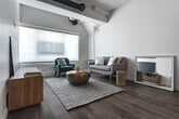 Located at 3550 W 6th St in the Koreatown neighborhood of Los Angeles, this single-floor unit blends modern design with urban practicality. The open-concept layout connects the kitchen and living areas seamlessly, creating a fluid and functional space. The kitchen is outfitted with a full suite of stainless steel appliances — including a refrigerator, range, microwave, and dishwasher — complemented by white cabinetry, light wood flooring, and a bar-height dining table that provides a casual spot for meals. The living area extends toward a large window that draws in natural light, featuring exposed structural beams and visible ductwork overhead that lend an industrial-loft character. Wood flooring continues throughout, adding warmth and cohesion. The bedroom is a calm, well-proportioned retreat with soft neutral walls, wood flooring, and ample space for furnishings. Adjacent to the bedroom, the bathroom is finished with large-format white tile, a combined tub and shower with a rainfall showerhead, and a wood-toned vanity with modern fixtures. The clean white walls and recessed lighting throughout the unit contribute to a bright, contemporary ambiance suited for urban living in Los Angeles.