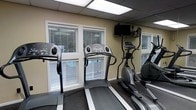 This single-floor fitness room is designed to provide a functional and comfortable workout environment. The space is outfitted with essential cardio equipment, including multiple treadmills and an elliptical trainer, arranged to maximize the available floor area. A rubberized flooring surface runs throughout, providing both safety and durability underfoot.

Natural light filters into the room through several large windows dressed with horizontal blinds, offering the option to control glare during workouts. The warm beige walls contribute to an inviting atmosphere, while a wall-mounted flat-screen television provides entertainment to help users stay engaged during their exercise sessions.

A large wall mirror on one side of the room visually expands the space and allows users to monitor their form. The drop ceiling with recessed panel lighting ensures the room is evenly and brightly illuminated. Overall, this fitness room delivers a clean, practical, and well-lit setting suited for daily cardiovascular training.