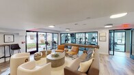 This single-floor amenity space is thoughtfully designed to serve as a social and wellness hub. The central lounge area sets a welcoming tone with light hardwood-style flooring, a neutral-toned sectional sofa, vibrant amber accent chairs, and a round textured coffee table atop a woven area rug. Large sliding glass doors flood the space with natural light and provide direct access to an outdoor patio with bar-height seating, seamlessly blending indoor and outdoor living.

A floor-to-ceiling glass partition wall visually connects the lounge to the fitness center, which is generously outfitted with treadmills, ellipticals, stationary bikes, a rowing machine, a dumbbell rack, a cable machine, and a wall-mounted television. The gym features rubber flooring, large windows on multiple walls that invite natural light from the surrounding landscape, and a distinctive circular blue pendant light fixture that adds a modern aesthetic touch.

The kitchenette is clean and functional, featuring white shaker-style cabinetry, a white subway tile backsplash, dark countertops, stainless steel appliances including a side-by-side refrigerator and dishwasher, and a built-in microwave. A water fountain is also accessible within the common area. Together, these spaces create a well-rounded amenity suite suited for both relaxation and an active lifestyle.