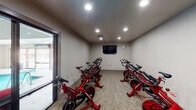 Located at 3751 Appian Way in Lexington, Kentucky, this dedicated spin studio offers a focused and functional environment for indoor cycling workouts. The single-room space is thoughtfully arranged to accommodate multiple red stationary spin bikes, positioned to give each rider ample space during individual or group cycling sessions. The room features neutral-toned walls and durable patterned carpeting underfoot, creating a practical yet comfortable exercise environment. Recessed lighting installed in the ceiling provides consistent, even illumination throughout the studio. A wall-mounted flat-screen television is centrally positioned at the front of the room, allowing riders to follow along with workout content or entertainment during sessions. A wall-mounted speaker accompanies the TV, enabling audio playback to enhance the cycling experience. The studio benefits from a large glass door and adjacent window that provide a direct visual connection to an outdoor pool area, allowing natural light to filter into the space. A secondary solid door offers an additional entry and exit point. The overall design of the studio is clean and utilitarian, keeping the focus squarely on fitness functionality. This space is well-suited for structured spin classes or individual cardio training within the broader amenity offerings of the property.