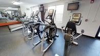 This fitness center is designed to support a comprehensive cardio workout experience within a compact, efficiently arranged space. The room is outfitted with multiple elliptical trainers, treadmills, and a recumbent stationary bike, providing users with a diverse range of low-impact and aerobic exercise options. The equipment is arranged to maximize floor space while maintaining comfortable clearance between machines. The flooring consists of durable, dark rubberized material, ideal for a high-traffic fitness environment and providing both safety and stability during workouts. Natural light filters in through slatted blinds on the windows, contributing to a bright and energizing atmosphere. The walls are finished in a clean, neutral white, keeping the space feeling open and uncluttered. A wall-mounted flat-screen television is positioned for easy viewing from most machines, offering entertainment during workouts. An additional monitor is also mounted near the equipment area, further enhancing the user experience. The ceiling is standard height, and ventilation is provided via a wall vent, ensuring adequate airflow. Overall, this fitness center offers a practical and well-maintained environment suited for daily cardiovascular exercise.
