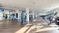 Located at 1213 Avalon Blvd in Alpharetta, GA, this fitness center offers a generously sized, single-floor exercise space designed for a full-spectrum workout experience. The room is bathed in natural light courtesy of large, black-framed windows that line multiple walls, with several overlooking an outdoor pool area—creating an energizing and visually open atmosphere.

The space features durable hardwood-style flooring throughout, crisp white walls, and recessed lighting complemented by ceiling fans that maintain comfortable airflow. Full-length mirrors along one wall enhance the sense of openness while providing functional visibility for strength training.

The facility is comprehensively equipped, including a row of treadmills and stationary bikes for cardio, stair climbers, ellipticals, and rowing machines. A dedicated free weights area offers a full dumbbell rack alongside adjustable benches, while multiple cable and selectorized strength machines support a variety of resistance training needs. Flat-screen TVs are mounted throughout the space for entertainment during workouts.

A storage and counter area near the entrance provides a convenient spot for personal items, and rubber mat flooring is placed strategically beneath strength equipment. The overall design strikes a clean, modern aesthetic that feels both functional and welcoming, making this facility a well-appointed amenity for users of all fitness levels.