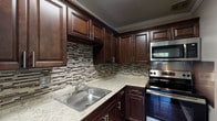 This single-floor unit offers a thoughtfully designed layout with a blend of modern updates and functional living spaces. The kitchen is a standout feature, showcasing rich dark-walnut cabinetry with brushed nickel hardware, light granite countertops, and a striking mosaic tile backsplash in neutral tones. Stainless steel appliances — including a range, over-the-range microwave, and dishwasher — complete the well-equipped cooking space, which also features white crown molding for a polished finish.

The living and bedroom areas are finished with warm, dark hardwood-style flooring that runs consistently throughout, providing a cohesive and inviting feel. Walls are painted in soft, muted tones of sage green and cool gray, creating a calm and neutral backdrop. The living area benefits from natural light through horizontal windows that offer views of surrounding greenery. A bedroom features the same cool-gray palette and hardwood flooring, offering a quiet and comfortable retreat.

The bathroom is clean and straightforward, featuring white subway tile surrounding a full soaking tub and shower combination, with a chrome towel bar and brushed nickel grab bar adding practical detail. The overall layout, as seen in the dollhouse view, is compact yet efficiently organized, with clear separation between living, sleeping, and kitchen areas.