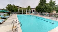 This outdoor aquatic facility centers around a generously sized rectangular swimming pool with vivid turquoise water, offering ample space for both lap swimming and leisure. The pool is accessed via stainless steel handrails and entry steps, ensuring safe and convenient entry for all users. A wide, light-toned concrete pool deck surrounds the water, providing plenty of room for lounging. Multiple green pool chairs and chaise lounges are arranged along the deck, offering comfortable spots for relaxation. The facility is enclosed by a metal perimeter fence, ensuring a secure environment. A dedicated pool house or clubhouse structure is visible at the rear of the space, likely housing restrooms or storage amenities. The surrounding landscape features a generous collection of mature deciduous trees, providing natural shade and a pleasant, park-like atmosphere. The overall setting is open and airy, with the tree canopy and neighboring structures creating a sense of privacy and seclusion. The space is well-suited for community recreation, combining functional swimming infrastructure with comfortable outdoor amenities.