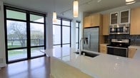 This single-floor space showcases a thoughtfully designed open-concept layout that seamlessly connects the kitchen and living areas. The kitchen serves as a focal point, centered around a generously sized island with a light-toned stone countertop, an undermount sink, and a contemporary arc faucet. Pendant lighting hangs above the island, providing both functional illumination and a design accent. Light maple cabinetry lines the perimeter, complemented by stainless steel appliances, including a French door refrigerator and dishwasher, creating a cohesive, modern aesthetic.

Warm hardwood flooring extends throughout, adding natural texture and continuity across the open plan. The living area is defined by an impressive wall of floor-to-ceiling windows with sleek black frames, flooding the interior with abundant natural light and offering views of the surrounding outdoor environment. The windows also provide access to what appears to be a balcony or outdoor area, further connecting the interior to the exterior. Track lighting and recessed ceiling fixtures supplement the natural light. The overall ambiance is clean, bright, and contemporary, suited to modern urban living.