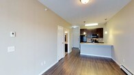 This single-floor residence offers a thoughtfully designed open-concept layout that seamlessly connects the living area to the kitchen. Rich hardwood flooring extends throughout the space, adding warmth and continuity to the overall aesthetic. The kitchen is a standout feature, showcasing dark espresso cabinetry that reaches toward the ceiling, light granite countertops, and a breakfast bar peninsula that provides both additional prep space and casual seating. Black appliances, including a refrigerator and range, complement the bold cabinetry, while a stylish pendant light fixture suspended above the counter adds a refined touch. The kitchen is also equipped with a stainless steel sink and additional task lighting overhead. The living area is defined by clean, neutral-toned walls and crisp white baseboards, creating a bright and inviting atmosphere. A doorway off the main corridor leads to a bathroom, visible with dark cabinetry finishes that mirror the kitchen's design language, ensuring a cohesive style throughout. The entry door is positioned along the rear corridor, providing a clear and functional flow through the space. Overall, the unit presents a clean, modern aesthetic with practical finishes suited for comfortable everyday living.