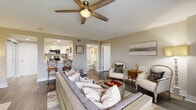 Located at 1200 South Courtenay Parkway on Merritt Island, Florida, this single-story residence offers a thoughtfully designed layout with one bedroom, two bathrooms, and a comfortable, efficient use of space. The open-concept living area is anchored by dark hardwood-style flooring, neutral warm-toned walls, and a ceiling fan that enhances airflow and comfort. Large sliding glass doors and windows flood the space with natural light and provide direct access to a private balcony with outdoor views. The kitchen features white shaker-style cabinetry, quartz countertops, stainless steel appliances, and a functional galley configuration that connects seamlessly to the living area via a breakfast bar. The spacious primary bedroom includes its own sliding glass door to the balcony, a ceiling fan, and generous closet space — including a walk-in closet with wire shelving. Both bathrooms are finished with clean, modern fixtures; one includes a walk-in shower with built-in niches and a quartz-topped vanity. Additional highlights include a dedicated utility room, a welcoming entrance, and a second closet for added storage. The overall ambiance is calm and contemporary, well-suited for comfortable living in the heart of Merritt Island.