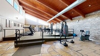 This fitness center combines striking architectural character with a comprehensive range of exercise equipment. The space is defined by its soaring vaulted ceiling with rich wood beams and exposed ductwork, creating an open and airy atmosphere. A row of clerestory windows along the upper walls floods the interior with natural light, enhancing the spacious feel throughout.

The layout is split into two distinct levels connected by steps and metal railings. The lower level features natural stone tile flooring and houses cardio equipment including stationary bikes, an elliptical trainer, and a treadmill, along with a punching bag station. A built-in wooden storage cabinet and framed artwork add a warm, welcoming touch to this area.

The raised upper level is carpeted and fully equipped for strength training, featuring a complete dumbbell rack, adjustable weight benches, cable machines, a multi-station strength system, and additional cardio machines including treadmills and a stair climber. Exposed brick walls line this section, contributing to the facility's rugged, energetic ambiance.

Wall-mounted sconces provide warm accent lighting, while mirrors strategically placed throughout the strength training area enhance the sense of depth. The overall design balances functional fitness infrastructure with architectural warmth, making it a distinctive and motivating environment for a full-body workout.