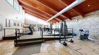 This fitness center combines striking architectural character with a comprehensive range of exercise equipment. The space is defined by its soaring vaulted ceiling with rich wood beams and exposed ductwork, creating an open and airy atmosphere. A row of clerestory windows along the upper walls floods the interior with natural light, enhancing the spacious feel throughout.

The layout is split into two distinct levels connected by steps and metal railings. The lower level features natural stone tile flooring and houses cardio equipment including stationary bikes, an elliptical trainer, and a treadmill, along with a punching bag station. A built-in wooden storage cabinet and framed artwork add a warm, welcoming touch to this area.

The raised upper level is carpeted and fully equipped for strength training, featuring a complete dumbbell rack, adjustable weight benches, cable machines, a multi-station strength system, and additional cardio machines including treadmills and a stair climber. Exposed brick walls line this section, contributing to the facility's rugged, energetic ambiance.

Wall-mounted sconces provide warm accent lighting, while mirrors strategically placed throughout the strength training area enhance the sense of depth. The overall design balances functional fitness infrastructure with architectural warmth, making it a distinctive and motivating environment for a full-body workout.