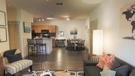 This single-floor residence showcases a thoughtfully designed open-concept layout that effortlessly connects the kitchen, dining, and living areas into one cohesive and functional living space. Rich, dark hardwood flooring flows throughout, anchoring the space with warmth and a contemporary aesthetic.

The kitchen is outfitted with warm honey-toned wood cabinetry, black appliances, and a breakfast bar with bar seating — ideal for casual dining or entertaining. Track lighting runs along the ceiling, providing flexible, directed illumination across both the kitchen and dining zones.

The dining area comfortably accommodates a full table and chair set, centered on a round area rug that defines the space. Adjacent to it, the living area offers a relaxed yet stylish setting with an upholstered sofa and bold accent pillows that add a pop of color to the neutral palette.

The walls are painted in a clean, warm neutral tone that enhances the sense of openness, while the open floor plan allows light and sightlines to travel freely from one end of the space to the other. A hallway visible from the living area suggests additional private rooms beyond. Overall, the space balances modern design sensibility with an inviting, livable atmosphere.