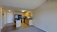 This single-floor residence offers a practical and inviting layout centered around an open-concept kitchen and adjoining living area. The kitchen is well-appointed with warm wood-toned cabinetry on both upper and lower levels, complemented by dark countertops that provide a clean contrast. Black appliances, including a range, microwave, and dishwasher, lend a cohesive and modern aesthetic to the cooking space. A peninsula with a white base extends into the room, offering additional prep space and a natural dividing point between the kitchen and the rest of the living area. The kitchen floor is finished in neutral-toned tile, which transitions smoothly into plush carpeting in the adjoining living space. Recessed and flush-mounted ceiling lighting keeps the kitchen well-lit, while the open layout allows light and space to flow freely between zones. The neutral wall tones throughout create a calm, versatile backdrop suitable for a variety of interior styles. The seamless connection between the kitchen and living area makes this floor plan well-suited for both everyday living and casual entertaining. Overall, the space communicates a functional, comfortable design with thoughtful attention to practicality and flow.