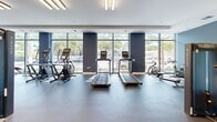 Located at 2920 Northwest 18th Avenue in Miami, FL, this fitness center offers a generously proportioned, single-floor exercise space designed to support a comprehensive workout experience. The facility is divided into two distinct functional zones that together accommodate a wide range of training needs.

The cardio area is bright and inviting, characterized by floor-to-ceiling windows along an entire wall that flood the space with natural light and provide open views of the surrounding greenery and streetscape. This zone houses multiple treadmills, elliptical trainers, and a rowing machine, all arranged to maximize the sense of openness. Navy blue accent columns punctuate the space, adding a modern, intentional design touch against the crisp white walls and light-colored rubber flooring.

The strength training area transitions to a darker, more focused atmosphere with speckled rubber flooring for durability and safety. It is equipped with professional-grade Matrix machines, a functional trainer cable system, a leg press, free weight benches, barbells, and a wall-mounted mirror that enhances the sense of depth. Recessed lighting provides consistent illumination throughout.

The facility features a clean, contemporary aesthetic with a neutral color palette, high ceilings, and double-door entry access. The thoughtful layout ensures ample open floor space between equipment, allowing for comfortable movement and a focused training environment.