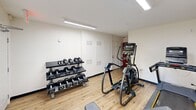 Located at 4515 Apex Hwy in Durham, NC, this fitness center offers a practical and well-equipped exercise space designed to support a variety of workout routines. The room features light wood-style flooring, white walls, and bright overhead fluorescent lighting that creates a clean, energizing atmosphere. Natural light filters in through a window fitted with horizontal blinds, adding a welcoming feel to the space. Climate comfort is provided by a wall-mounted HVAC unit positioned beneath the window. The exercise room is furnished with a Matrix elliptical machine and a treadmill for cardiovascular training, alongside an adjustable workout bench for strength and flexibility exercises. A multi-tier dumbbell rack holds a full range of free weights, making the space suitable for both light toning and more intensive strength training. A bulletin board is mounted on the wall, serving as a functional informational area. A row of bold red metal lockers provides secure personal storage for users. The room is equipped with a security camera and clearly marked emergency exit signage, ensuring a safe environment. Two access doors offer convenient entry and exit points. Overall, this fitness center delivers a compact yet fully functional workout facility.