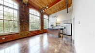 This single-floor loft showcases a masterful blend of historic industrial character and modern finishes. The open living area commands attention with its soaring wood-beamed ceiling, exposed ductwork, and original brick walls punctuated by tall, arched multi-pane windows that draw in abundant natural light and offer lush outdoor views. Polished concrete floors run throughout, reflecting light and anchoring the space with an urban, contemporary feel.

The kitchen is well-appointed with shaker-style cabinetry, dark stone countertops, stainless steel appliances, a decorative tile backsplash, and a substantial central island with pendant lighting overhead — all framed against the backdrop of the iconic brick wall and oversized windows. A secondary kitchen or utility area features additional cabinetry and a striking patterned tile backsplash with open shelving.

The bedroom is generously proportioned with high ceilings and access to a spacious walk-in closet with wire shelving. The bathroom is finished with a dual-sink vanity with dark countertops, round mirrors, brushed nickel fixtures, and a full tiled tub-shower surround. Track lighting, ceiling fans, and wall sconces add practical and stylish illumination throughout this thoughtfully designed industrial loft space.
