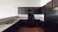 This compact yet well-designed apartment in Detroit offers 550 square feet of contemporary living space. The single-level floor plan includes a spacious 180 square foot bedroom, a full bathroom, and an open-concept living area. The kitchen is a standout feature, boasting dark wood cabinetry, granite countertops, and black appliances, creating a striking contrast against the light walls. A breakfast bar with a granite overhang provides additional seating and separates the kitchen from the living area. Throughout the apartment, warm-toned hardwood floors add a touch of elegance and continuity. The bedroom features large windows that allow ample natural light, complemented by recessed lighting fixtures. The bathroom, while compact, is efficiently designed with modern fixtures. A hallway connects the various spaces, likely housing additional storage options. The apartment's neutral color palette and clean lines create a blank canvas for personalization, making it suitable for a variety of tastes and lifestyles.