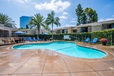 Located at 15555 Huntington Village Lane in Huntington Beach, California, this two-story residential community embodies the relaxed, sun-soaked lifestyle of Southern California. The property's architecture features a warm stucco exterior with balconies and large windows that provide residents with views of the beautifully maintained grounds below.

The centerpiece of the outdoor amenity space is a generously sized, freeform swimming pool with crystal-clear turquoise water. A striking natural rock waterfall feature anchors one end of the pool area, adding a resort-like aesthetic to the environment. The expansive pool deck is appointed with blue lounge chairs and shaded umbrella seating areas, offering ample space for sunbathing and relaxation under the California sun.

Towering palm trees and mature landscaping frame the pool courtyard, creating a lush, tropical ambiance throughout the outdoor space. Manicured green lawn areas and vibrant flowering plants add color and texture to the surroundings. The pool area is enclosed by a wrought iron fence, providing both security and a sense of privacy for residents. A dedicated sport court is also visible on the property, further enhancing the range of recreational amenities available. The overall setting reflects a well-maintained community designed for comfortable outdoor living in the heart of Huntington Beach.