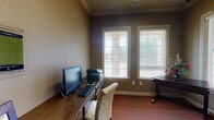 This single-floor space is thoughtfully designed to serve as both a functional office and a creative activity room. The interior is finished with warm taupe and deep gray-brown wall tones, complemented by elegant crown molding that adds a refined architectural detail throughout. Rich hardwood-style flooring transitions into textured, multi-toned carpeting, grounding the space with warmth and comfort underfoot.

The room is divided into two distinct functional zones. One side features a dedicated computer workstation with a built-in desk, providing ample surface space for a desktop monitor, keyboard, and printer, along with an upholstered chair for comfortable use. The opposite side offers a secondary wooden desk and additional cabinetry with drawers and cabinet doors, ideal for organizing craft or activity supplies. Wall-mounted horizontal rails provide convenient display and storage for rolled materials such as wrapping paper or craft sheets.

Multiple windows fitted with white horizontal blinds line the walls, allowing generous natural light to filter into the space while offering views of the surrounding greenery outdoors. A striking large decorative indoor tree adds a lively, natural element to the room's ambiance. A glass-paned interior door with a classic divided-light design connects the space to the adjoining area, enhancing the open and airy feel of this versatile, well-appointed room.