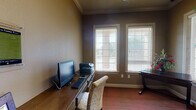 This single-floor space is thoughtfully designed to serve as both a functional office and a creative activity room. The interior is finished with warm taupe and deep gray-brown wall tones, complemented by elegant crown molding that adds a refined architectural detail throughout. Rich hardwood-style flooring transitions into textured, multi-toned carpeting, grounding the space with warmth and comfort underfoot.

The room is divided into two distinct functional zones. One side features a dedicated computer workstation with a built-in desk, providing ample surface space for a desktop monitor, keyboard, and printer, along with an upholstered chair for comfortable use. The opposite side offers a secondary wooden desk and additional cabinetry with drawers and cabinet doors, ideal for organizing craft or activity supplies. Wall-mounted horizontal rails provide convenient display and storage for rolled materials such as wrapping paper or craft sheets.

Multiple windows fitted with white horizontal blinds line the walls, allowing generous natural light to filter into the space while offering views of the surrounding greenery outdoors. A striking large decorative indoor tree adds a lively, natural element to the room's ambiance. A glass-paned interior door with a classic divided-light design connects the space to the adjoining area, enhancing the open and airy feel of this versatile, well-appointed room.