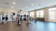 Located at 5401 Commonwealth Centre Pkwy in Midlothian, VA, this fitness center offers a well-appointed, single-floor space dedicated to health and exercise. The room features durable rubber flooring throughout, providing a safe and practical surface for a full range of workouts. Recessed lighting illuminates the space evenly, complemented by abundant natural light streaming through multiple large windows with views to the outdoors. The cardio zone includes treadmills and an elliptical trainer, each positioned alongside wall-mounted mirrors that enhance the sense of openness. A dedicated strength training area houses a multi-station cable machine, a weight bench, and a full dumbbell rack with a wide range of weights. Additional Hoist-brand strength machines are arranged throughout the room to support targeted muscle training. One wall features a bold navy accent with a wall-mounted flat-screen television and a built-in shelving unit, adding a functional and stylish focal point. Storage cubbies, medicine balls, and a water station round out the amenities. The overall ambiance is clean, modern, and motivating, making this fitness center a fully equipped destination for residents seeking a convenient on-site exercise facility.