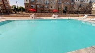 This outdoor pool area offers a refreshing retreat defined by its generously sized swimming pool filled with clear, turquoise water. The pool's smooth surface reflects the surrounding environment, creating a bright and inviting atmosphere on sunny days. A wide concrete pool deck surrounds the water, providing ample space for movement and relaxation. Along the poolside, white patio tables and chairs are arranged beneath vibrant red umbrellas, offering shaded seating areas ideal for lounging or socializing. Entry to the pool is facilitated by built-in stainless steel handrail ladders. The area is enclosed by a classic black wrought iron fence, ensuring both security and an open, airy feel that maintains visibility to the surroundings. A low brick retaining wall runs along the back edge of the deck, adding a structured, architectural boundary to the space. The backdrop features an attractive multi-story building with brick and neutral-toned façades, complementing the overall aesthetic. The pool area is well-maintained and thoughtfully designed to provide a comfortable and functional outdoor amenity for residents.