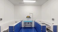 Located at 28 Southfield Ave in Stamford, CT, this single-floor professional dog wash facility is purpose-built for efficient and thorough pet grooming. The space is designed with cleanliness and functionality at its core, featuring bright white paneled walls and a vibrant blue speckled epoxy floor that is both durable and easy to sanitize.

The main grooming area is equipped with two raised bathing tubs, each accented with bold blue cabinetry and fitted with plumbing fixtures including a handheld spray attachment. Centrally positioned between the tubs is a light blue grooming table with a sturdy metal frame and an overhead grooming arm mount, accompanied by a professional-grade blow dryer — providing a complete grooming station in one compact area.

Overhead fluorescent lighting ensures the entire space is well-lit, while ceiling-mounted ventilation supports a comfortable working environment. The facility is accessed through a set of full-height glass sliding doors, offering visibility into the adjacent parking structure and giving the space an open, accessible feel. A textured entry mat at the entrance adds a practical touch.

Additional features include a built-in step or ramp surface for easy pet access to the tubs, smooth white wall panels for hygienic maintenance, and well-organized utility connections. The overall layout is thoughtfully designed to accommodate both pets and groomers with ease and efficiency.