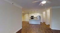 This single-floor residence showcases an open-concept layout that seamlessly connects the living area to the kitchen, creating a spacious and inviting atmosphere. Rich hardwood flooring runs throughout, complementing the warm, neutral wall tones and crisp white trim accents.

The kitchen is a standout feature, equipped with stainless steel appliances including a refrigerator and microwave. White shaker-style cabinetry extends to the upper walls, providing ample storage, while a center island with a light stone countertop offers additional workspace and a casual dining option. Elegant pendant lights hang above the island, adding a refined touch, and under-cabinet task lighting enhances functionality.

Decorative wainscoting lines the lower walls of the kitchen and living area transition, contributing a classic architectural detail. Crown molding runs along the ceiling perimeter, elevating the overall finish of the space. A ceiling fan in the living area ensures comfortable airflow year-round. Track lighting in the living space provides flexible illumination. The open layout and cohesive design elements give the home a clean, modern aesthetic with traditional detailing throughout.