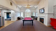 Located at 4122 Avalon Dr in Peabody, MA, this single-floor clubhouse space is a well-appointed gathering destination designed for both relaxation and entertainment. The living room is anchored by soaring ceilings with coffered detailing, a classic fireplace framed in white millwork, and an elegant arched entryway. A sweeping arched transom window floods the space with natural light, while French doors open directly to an outdoor area, seamlessly connecting interior and exterior. Sophisticated cloud-motif wallpaper lines the walls alongside crisp wainscoting, and a bold patterned carpet grounds the room. A red billiards table serves as a striking centerpiece, complemented by a wall-mounted television and curated seating. The dining room transitions into a vibrant game room, distinguished by deep charcoal walls beneath a dramatic orange ceiling. A round poker table sits at the heart of the room, surrounded by sleek black leather chairs. Wall-mounted Scrabble and tic-tac-toe boards add a playful, interactive dimension to the space. Accent tables in red add pops of color throughout, while patterned carpet and a statement pendant light complete the bold, curated aesthetic of this dynamic and inviting space.