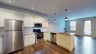 Located at 122 Parsons Ave in Columbus, Ohio, this single-floor unit offers a thoughtfully designed open-concept layout that seamlessly connects the kitchen and living areas. Rich, dark hardwood flooring runs throughout the main living spaces, providing a warm and grounding contrast to the light blue-gray walls and crisp white trim.

The kitchen is a standout feature, outfitted with white shaker-style cabinetry, a classic white subway tile backsplash, and a full suite of stainless steel appliances including a refrigerator, range, microwave, and dishwasher. A peninsula with a light-toned countertop and an integrated sink extends into the living area, offering additional prep space and casual seating. Pendant lighting in an industrial black finish hangs above the peninsula, complementing the track lighting overhead.

The open living area benefits from natural light provided by windows near the entry door, creating an airy and welcoming ambiance. A walk-in closet with wire shelving and plush carpeting offers generous storage. The bathroom features large-format gray tile flooring, a walk-in shower with white subway tile surround, a glass panel door, and matte black fixtures that add a sleek, modern touch. The overall design balances modern aesthetics with functional, everyday living.
