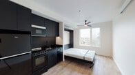 This thoughtfully designed studio unit at 5620 N Interstate Ave in Portland, Oregon, offers a clean, modern aesthetic through a carefully curated combination of form and function. The open-plan living area features light hardwood flooring that runs throughout, creating a warm contrast to the bold design elements. A built-in Murphy bed folds seamlessly against a matte black accent wall, maximizing usable floor space while maintaining a contemporary look. A ceiling fan with integrated lighting anchors the living area, which is filled with natural light from a large picture window overlooking the surrounding Portland neighborhood. The kitchen is outfitted with floor-to-ceiling matte black cabinetry, a black gas range, an under-cabinet microwave, and dark countertops — all working together to create a striking and cohesive culinary space. The private bathroom features wood-look tile flooring, white subway tile in the walk-in shower, matte black hardware accents, and a wall-mounted sink, delivering a clean and modern finish. The unit's neutral white walls and efficient single-floor layout create a cohesive, minimal ambiance that balances style and practicality in one of Portland's well-connected neighborhoods along North Interstate Avenue.