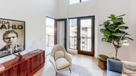 This thoughtfully designed two-floor residence showcases a blend of modern aesthetics and functional living across a well-organized layout. The main living area impresses with soaring double-height ceilings and an abundance of natural light, creating an open, airy ambiance. Large black-framed French doors open directly onto a private balcony, offering sweeping urban skyline views.

The kitchen is a standout space, featuring two-tone cabinetry with navy lower cabinets and white upper cabinets, complemented by a textured white tile backsplash, brass fixtures, a deep undermount sink, and a full suite of stainless steel appliances. Light hardwood flooring flows consistently throughout the residence, lending warmth and continuity.

The bedrooms are bright and well-proportioned. The primary bedroom offers balcony access through a large picture window with city views, while additional bedrooms provide versatile spaces suitable for sleeping or use as a home office. The upper floor includes a dedicated office or flex area positioned at the top of the staircase, ideal for focused work.

The bathroom is finished with classic white subway and hexagonal floor tiles, a glass-enclosed walk-in shower, and matte black and brass hardware accents. A loft-style dollhouse view reveals the efficient, multi-room floor plan that maximizes every level of this well-appointed urban residence.